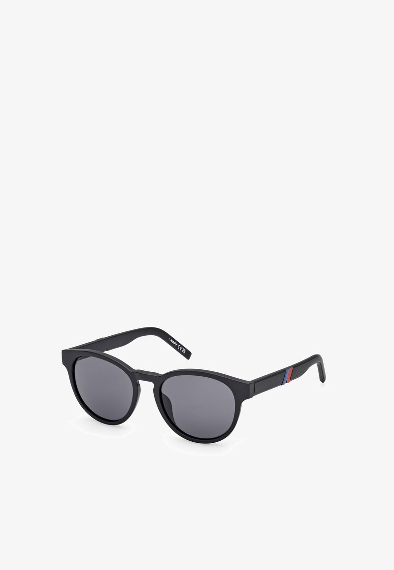 Schwarze Sonnenbrille mit runden Gläsern, mattem Finish, dicken Rahmen und seitlichen Akzenten in Rot und Blau. Die Gläser sind dunkel und polarisiert.
