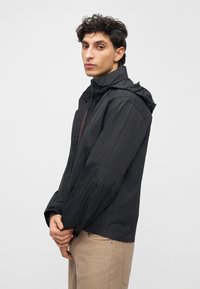 Casaco preto leve com capuz, apresenta um fecho de correr frontal e tecido texturizado. Tem um corte casual e é combinado com calças bege.