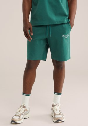 Mand iført grønne shorts med teksten "Leisure Club", hvide sokker med grønne striber og hvide løbesko, stående mod en ensfarvet baggrund.