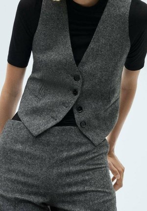 Personne portant un gilet gris boutonné et un pantalon assorti sur un haut noir à manches courtes, une main reposant près de la hanche.