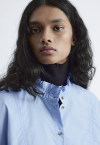 Light blue collared shirt with a ruffled neckline and brown button detail, worn over a black high-neck top. Soft fabric texture.