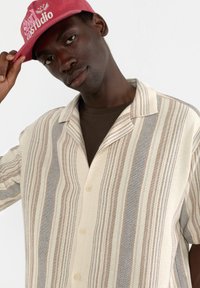 Chemise à manches courtes rayée en crème, marron et gris, fabriquée en tissu texturé. Présente un col à revers et une patte de boutonnage à cinq boutons.