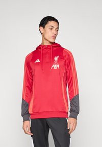 Červená mikina s kapucí Liverpool FC se �šedými doplňky, vybavená předním zipem, nastavitelnou kapucí a pruhy podél ramen. Zobrazeno logo Adidas.