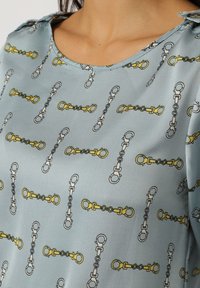 Blusa azul clara con un patrón de cadena repetido en amarillo, gris y negro, con un escote amplio y un ligero detalle en los hombros.