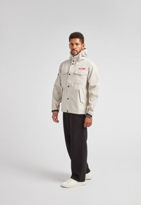 Lys grå vandtæt jakke med flere lommer, sikret med sorte knapper. Har en hætte og en kontrastfarvet rød label. Sorte bukser og hvide sko.