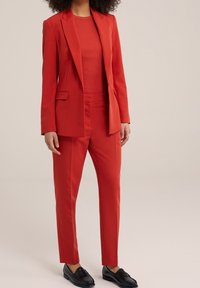 Červený oblek na míru, který obsahuje sako na jedno knoflík se dvěma předními kapsami a úzké kalhoty, doplněné o ladící top a černé mokasíny.