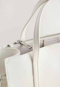 Weiße Lederhandtasche mit strukturierter Oberfläche, ausgestattet mit silbernen Reißverschlussbeschlägen, zwei kurzen Griffen und einem geräumigen Innenraum.