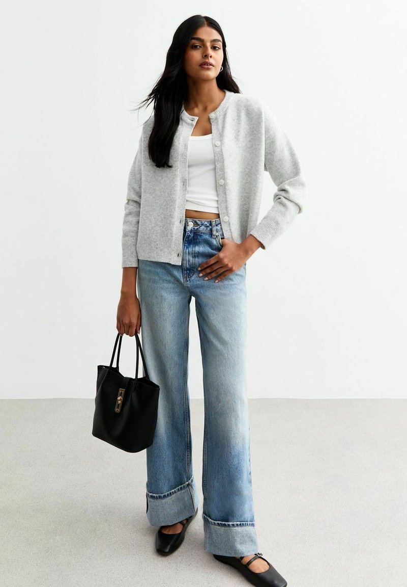 Cardigan grigio chiaro, canotta bianca aderente, jeans a gamba larga blu chiaro con orlo sfilacciato, borsa a spalla nera e scarpe nere basse con cinturini.