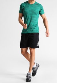 Zelené pruhované sportovní tričko z prodyšného materiálu, v kombinaci s černými šortkami s bílým logem a šedými sportovními botami.