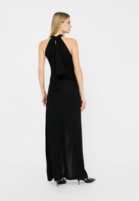 Zwarte maxi-jurk met een halternek en een gestrikt achterdetail. Glad, vloeiend stof met een subtiele glans. Ggedragen met zwarte hoge hakken.