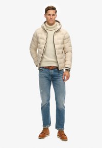 Mand iført en beige pufferjakke over en creme farvet turtleneck sweater, blå jeans med brunt bælte og brune læder støvler, stående mod en hvid baggrund.