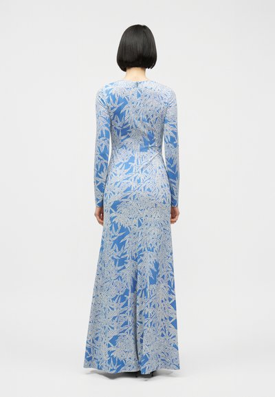 Γυναίκα με κοντά μαύρα μαλλιά που φοράει μπλε μακρυμάνικο φόρεμα μέχρι το πάτωμα με λευκά φυλλώδη σχέδια, όρθια με την πλάτη προς τα εμπρός σε λευκό φόντο.