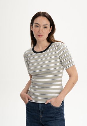Junge Frau mit langen braunen Haaren, die ein kurzärmeliges, beige-weiß gestreiftes Shirt und dunkelblaue Jeans trägt, steht mit den Händen in den Taschen.