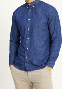 Mężczyzna w dopasowanej, długorękawowej, niebieskiej koszuli z denimu z guzikami, beżowych spodniach, z rękami w kieszeniach, stojący na jednolitym tle.