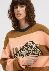 Padded leopard-print clutch in cream with black accents, featuring a top zip closure and logo detail. Model wears a brown and pink sweater.