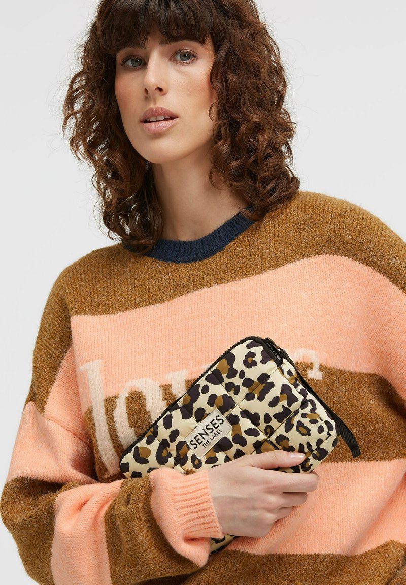 Padded leopard-print clutch in cream with black accents, featuring a top zip closure and logo detail. Model wears a brown and pink sweater.