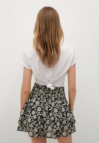 Femme aux longs cheveux châtain clair portant un t-shirt blanc noué et une jupe noire à fleurs superposée, tournée de dos contre un fond uni.