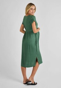 Groene gemusterde zwangerschapsjurk met korte mouwen, een geplooide taille en een zijsplit. Soepele textuur met een knie-lange snit. Subtiele geometrische print.