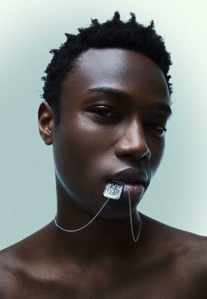 Rövid, göndör hajjal rendelkező fiatal személy díszes ezüst medálos nyakláncot tart az ajkai között, egyszínű világos háttér előtt.