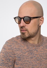 Ochelari de soare rotunzi cu rame negre și lentile maronii cu degradé. Haina este un pulover tricotat texturat, de culoare portocalie și gri.