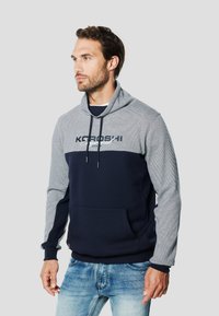 Sudadera con capucha que presenta un cuerpo azul marino con mangas a rayas grises, cordones ajustables y un bolsillo frontal tipo canguro.