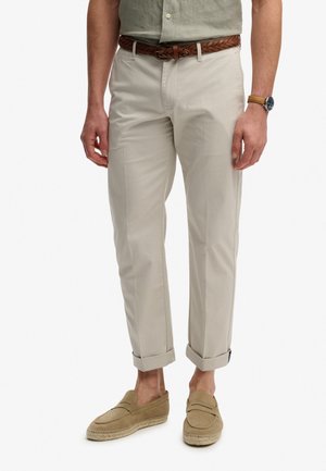 Pantalones chinos beige con corte de pierna recta, puños enrollados y textura sutil, combinados con un cinturón trenzado marrón y mocasines marrón claro.