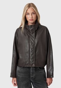 Tumši brūns ādas jaciņa ar augstu apkakli, saīsinātu dizainu un smalku spīdumu. Tam ir brīvi piedurknes un nav redzamu aizdari.