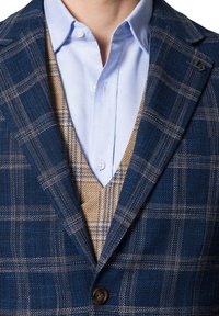 Niebieska marynarka w kratę z teksturowanej tkaniny, z brązowymi i białymi akcentami wzorów. Nałożona na beżowy kamizelkę i jasnoniebieską koszulę z kołnierzykiem.