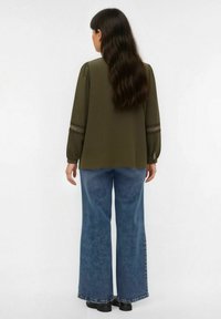 Olijfgroene blouse met lange mouwen en pofmouwen, afgewerkt met kant, gecombineerd met brede spijkerbroeken in blauw en zwarte schoenen, gezien van achteren.