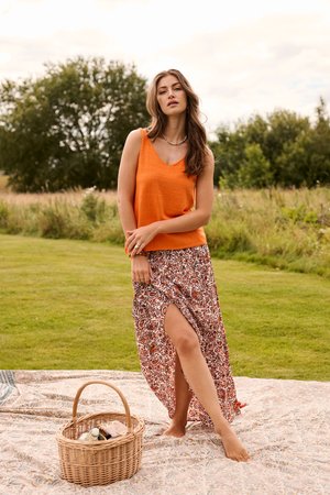 Naine oranži varrukateta topi ja lillelise lõhikuga seelikuga, paljajalu teki peal seismas, pikniku korvi lähedal rohelises õues.