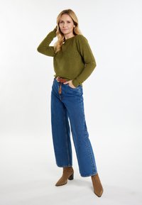 Maglione verde oliva con tessuto testurizzato, abbinato a jeans blu a vita alta e stivali alla caviglia marroni. Il modello posa con una mano tra i capelli.