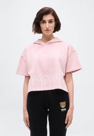 Kobieta ubrana w różową krótką bluzę z kapturem z napisem "Moschino" oraz czarne spodnie z logo misia i napisem "Moschino Underbear".