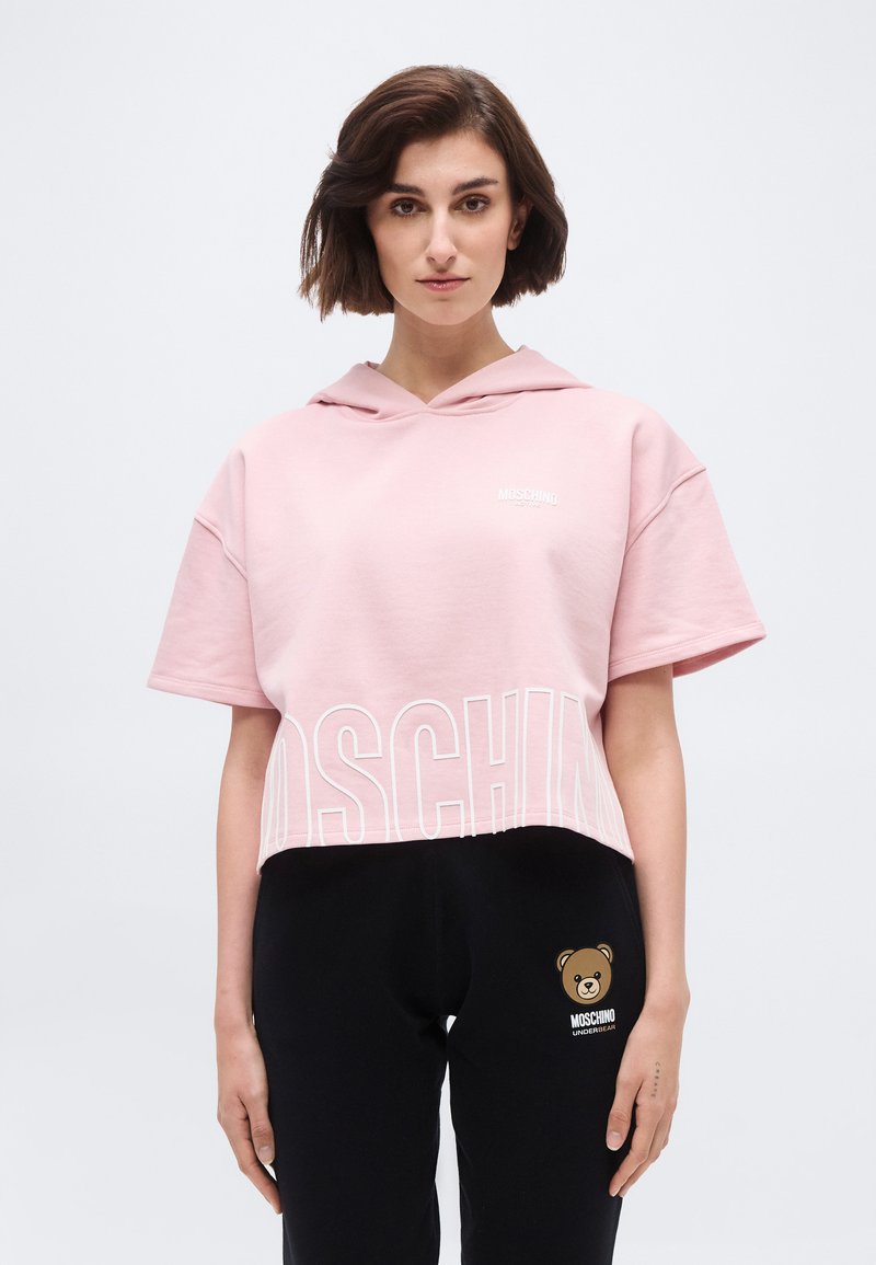 Woman wearing a pink cropped hoodie with "Moschino" text and black pants featuring a teddy bear logo and "Moschino Underbear" text.