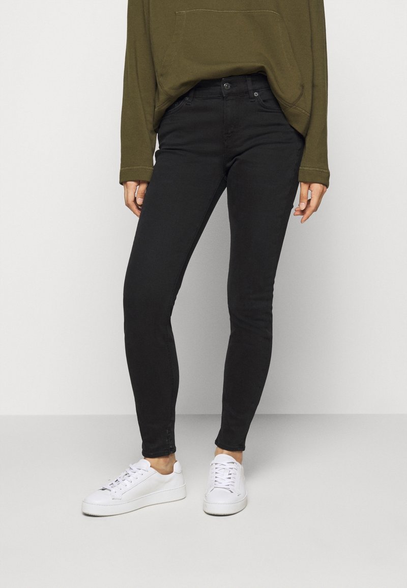 Jean skinny noir avec une texture lisse, dotés de poches avant et d'une coupe slim, associés à des baskets blanches et à un haut oversize vert olive.