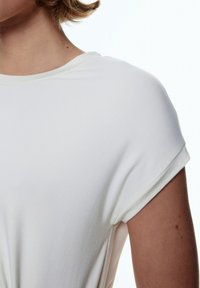 Weißes Baumwoll-T-Shirt mit rundem Halsausschnitt und umgeschlagenen kurzen Ärmeln. Angenehme Textur und lockere Passform, die subtile Nähdetails präsentiert.