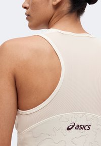 Gros plan d'une personne portant un débardeur beige Asics en tissu mesh avec des motifs abstraits cousus dans le dos.
