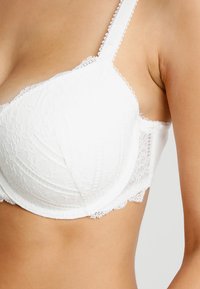 White lace bra featuring molded cups, intricate floral patterns, scalloped edges, and adjustable straps. Smooth fabric with detailed texture.
