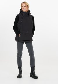 En isoleret sort vest med høj krave, lynlås foran og en stor lomme på fronten. Båret over en langærmet sort bluse og grå jeans.
