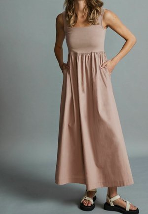 Femme portant une robe longue beige sans manches avec des poches et des sandales de couleur claire sur un fond gris uni.