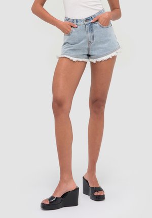 Vrouw draagt lichtblauwe denimshorts met rafelige zoom en zwarte plateau sleehak sandalen, staat met handen in de zakken.