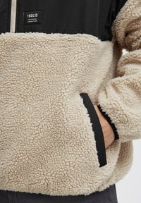 Pullover in fleece con corpo texturizzato beige, parte superiore nera e zip. Presenta tasche con rifiniture nere e un'etichetta sul petto.