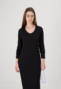Vestido longo preto com mangas compridas e um design de decote em V profundo, silhueta ajustada e textura suave; combinado com uma pequena bolsa roxa.