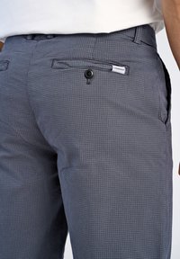 Pantaloni a scacchi grigi con un tessuto testurizzato, caratterizzati da una chiusura con bottone e una tasca applicata. È visibile un'etichetta contrassegnata con "Lindbergh".