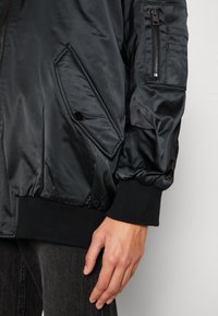 Veste bomber noire en satin avec poignets côtelés, dotée d'une poche zippée sur la manche et d'une poche à rabat boutonnée sur le devant.
