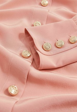 Camisa rosa claro con una textura de tela suave, adornada con seis botones dorados a lo largo de la tapeta y un cuello doblado.