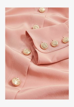 Heledad roosa särk, millel on sile kangatekstuur, kuue dekoratiivse kuldse nupuga nööbirea ääres ja voltitud krae.