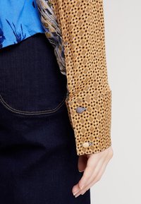 Dlouhý rukáv top s hnědým puntíkovaným materiálem, doplněný modrými knoflíky na manžetách, spojený s živou modrou květinovou vzorovanou částí.