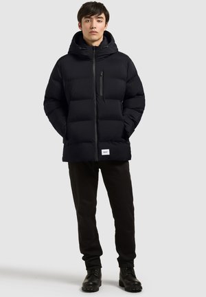 Jeune homme debout les mains dans les poches, portant une veste noire matelassée à capuche, un pantalon noir et des bottes noires sur un fond clair uni.