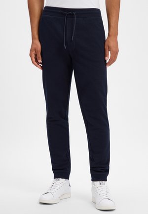 Man draagt donkerblauwe joggingsbroek met trekkoord in de taille en witte sneakers en staat op een effen achtergrond.