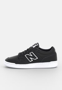 Baskets basses noires New Balance avec semelle blanche, logo blanc « N » sur le côté et lacets noirs sur un fond gris uni.